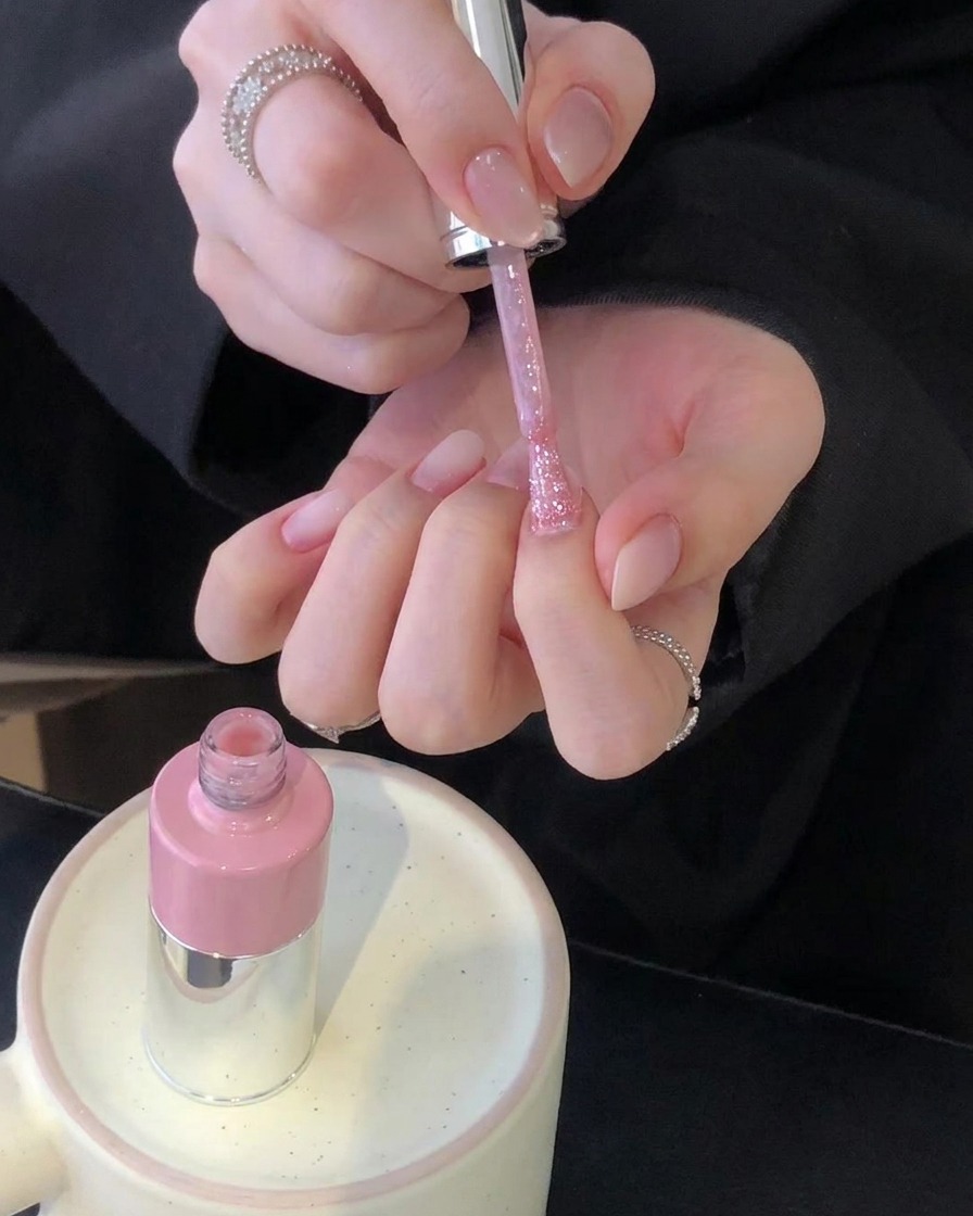 Una coppia di mani che tiene un pennello da smalto rosa sopra un set di unghie curate con un flacone rosa poggiato su un supporto in ceramica.