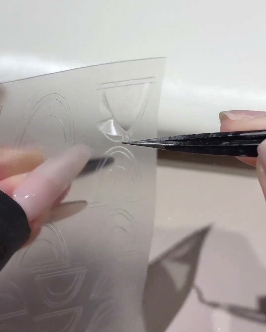 Un paio di pinzette che sollevano un adesivo guida French curvo da un foglio di plastica trasparente.