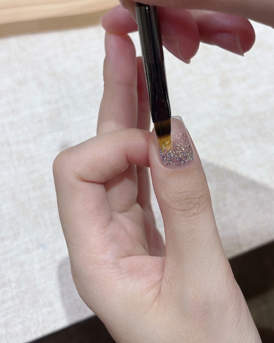 Una vista in primo piano di un pennello di dettaglio fine che applica uno strato di gel trasparente su una base per unghie con glitter scintillanti.