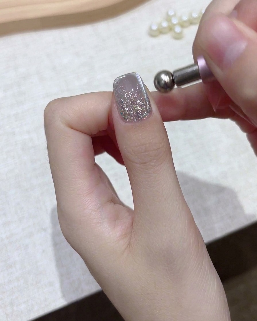 Un primo piano di una mano che tiene uno strumento magnetico sferico sopra unghie in gel glitterate argentate.