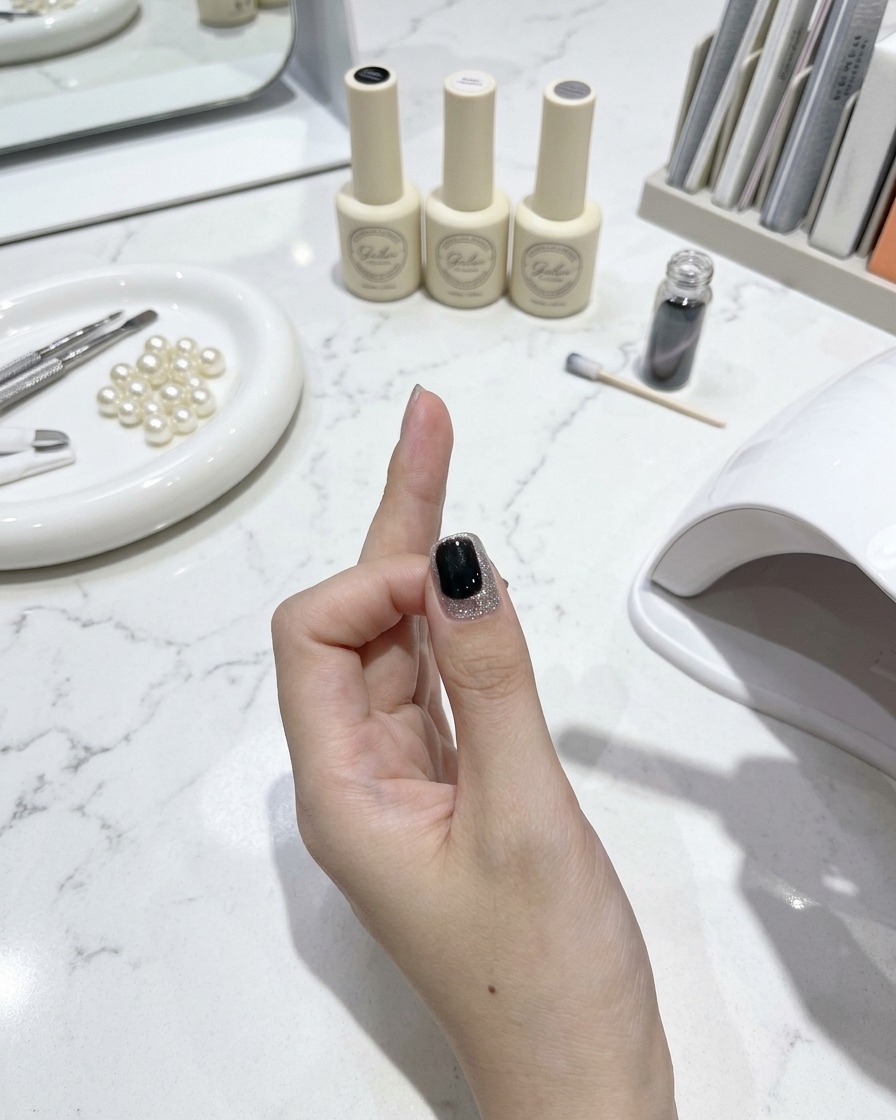 Un pennello per unghie che applica un top coat lucido su ununghia cat-eye finita con strumenti per manicure e boccette di smalto su un tavolo di marmo bianco sullo sfondo.