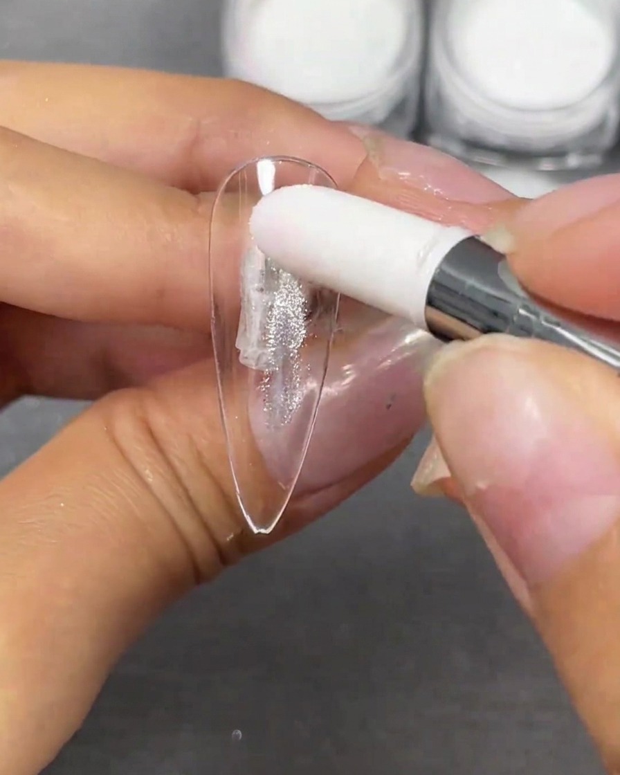 Una mano che utilizza un applicatore in spugna bianca per tamponare la polvere argentata Crystal Plating su una punta per unghie a stiletto trasparente.