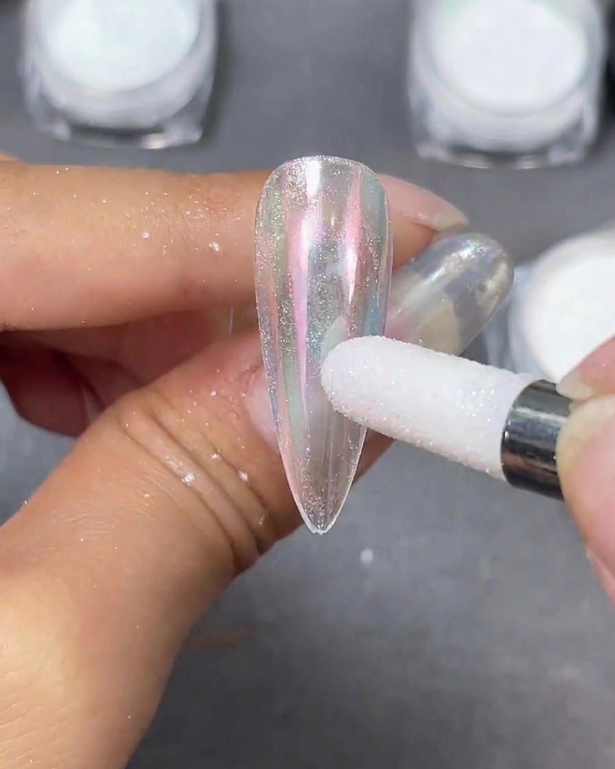 Un applicatore in spugna bianca che lucida la polvere perlata Moonlight su una punta per unghie a stiletto trasparente, creando una sfumatura iridescente.