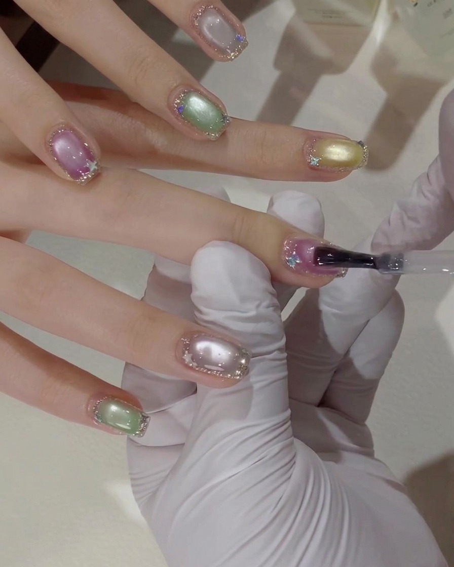 Primo piano di una mano con guanto che usa un pennello per stendere uno strato di top coat trasparente su ununghia cat eye rosa e viola con bordi glitterati argentati.