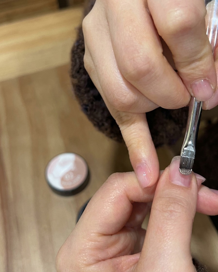 Primo piano di un pennello per gel che applica uno smalto scintillante oro nude su ununghia naturale.
