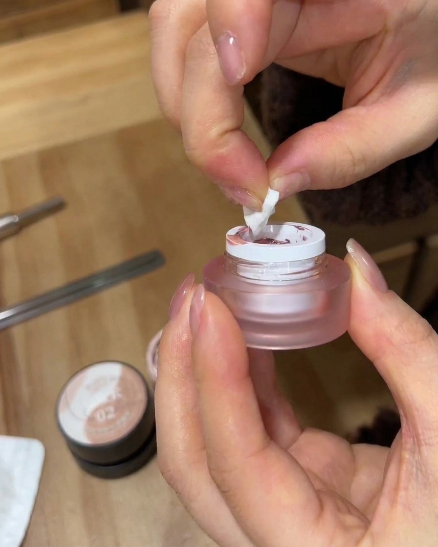Primo piano di mani che usano un pezzo strappato di spugnetta da trucco bianca per prelevare lo smalto gel rosa da un piccolo barattolo satinato.