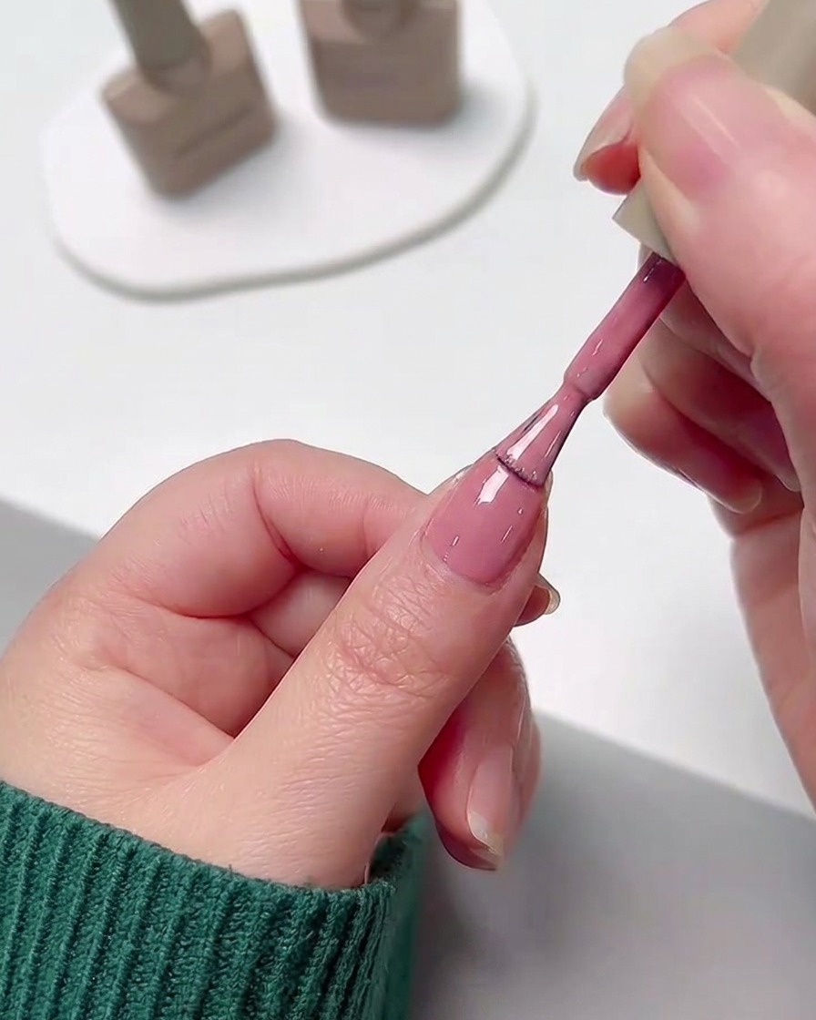 Una mano che applica uno strato uniforme di smalto gel rosa su ununghia usando il pennello del flacone.