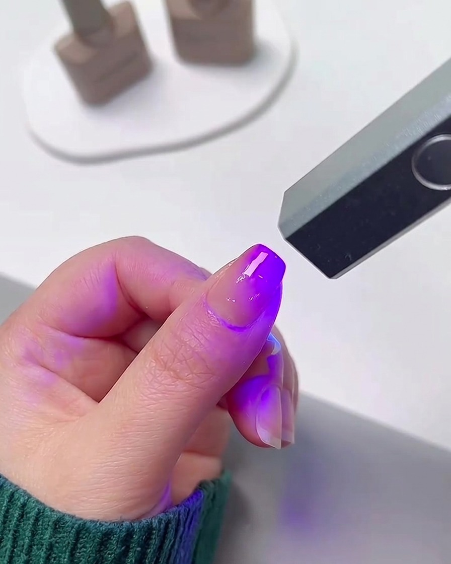 Un dito posizionato sotto una piccola lampada UV/LED portatile per unghie che emette una brillante luce viola.