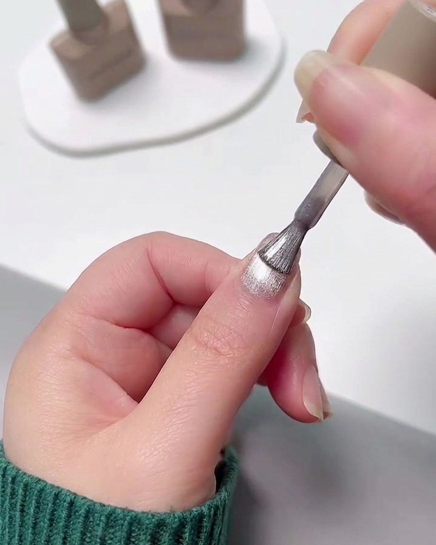 Una mano che applica uno strato di smalto gel cat eye argentato brillante sopra ununghia con sfumatura rosa polimerizzata.