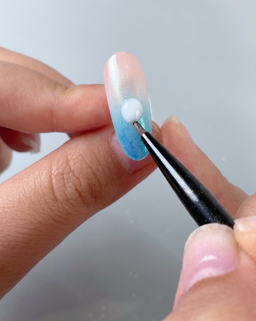 Un pennello fine per nail art che applica una goccia di gel autolivellante bianco latte in diagonale su ununghia sfumata.