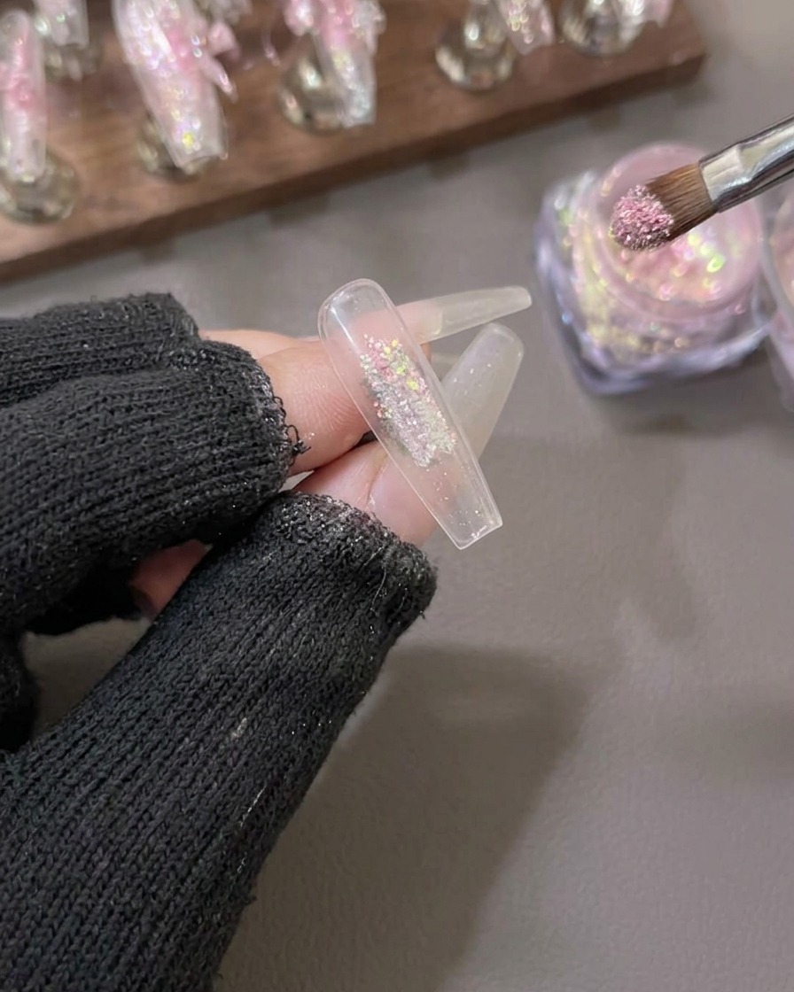 Un pennello applica polvere di scaglie aurora rosa e oro al centro di una tip per unghie trasparente.