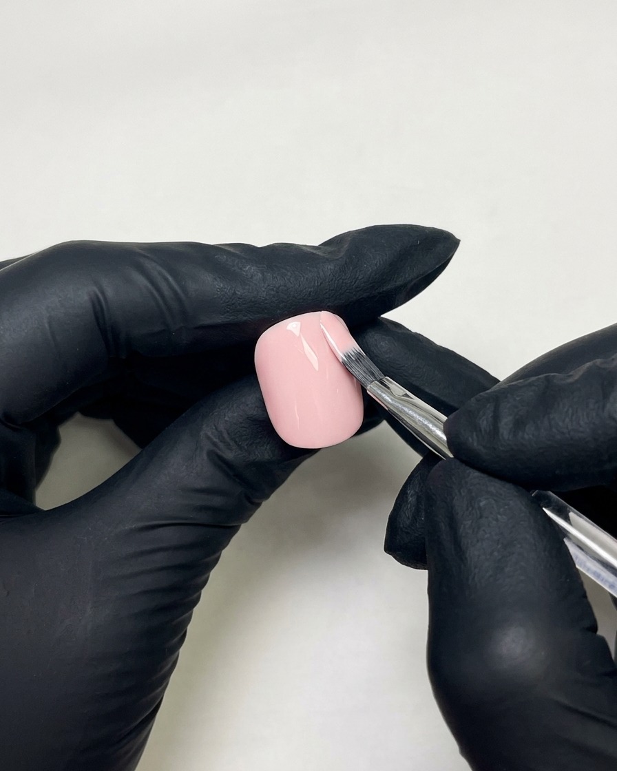 Mani con guanti che usano un pennello piatto per unghie per applicare lo smalto gel rosa carne su ununghia preparata.