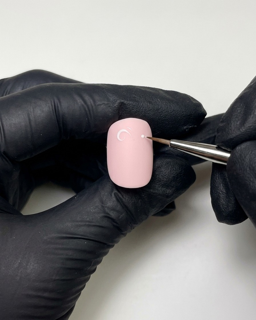 Mani con guanti che usano un pennello a punta fine con gel costruttore per disegnare un anello e un punto bianco-rosa su ununghia dipinta.