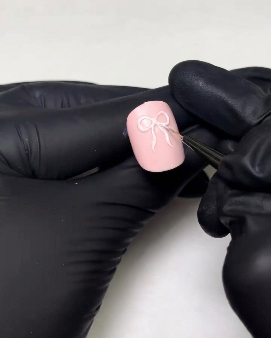 Una mano con guanto che utilizza un pennello da dettaglio fine per disegnare i passanti e le code del nastro di un design con fiocco 3D su ununghia rosa.