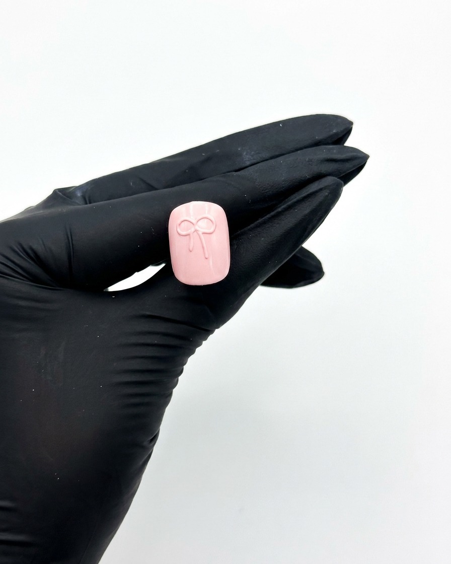 Una mano con guanto che tiene ununghia press-on rosa sotto una lampada per unghie UV per polimerizzare il design del fiocco 3D.