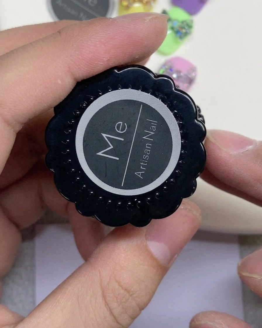 Una mano che tiene un barattolino nero con etichetta Artisan Nail e un logo circolare bianco.