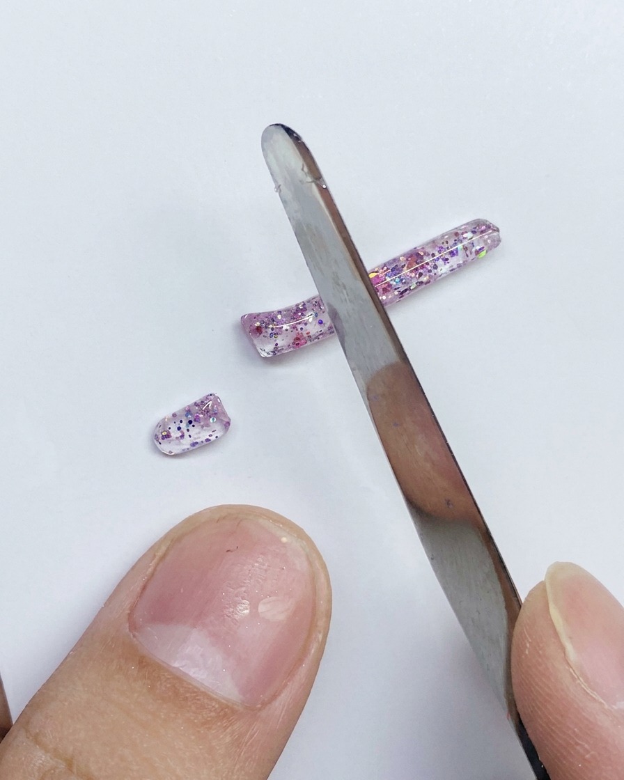 Una spatola di metallo che taglia in segmenti più piccoli una lunga striscia di gel PVC rosa glitterato su una superficie bianca.