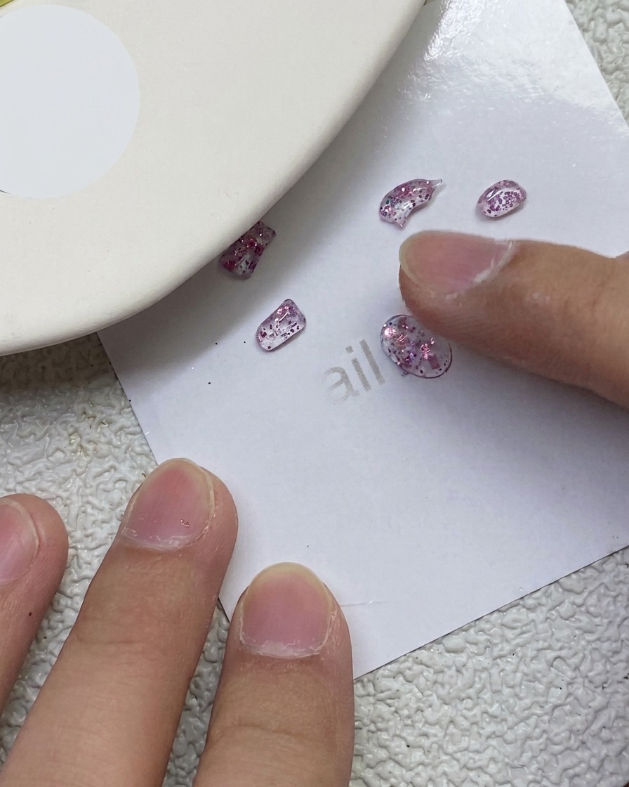 Un dito che preme e modella piccoli pezzi di gel PVC rosa glitterato a forma di fiocco su un foglio di carta bianco.