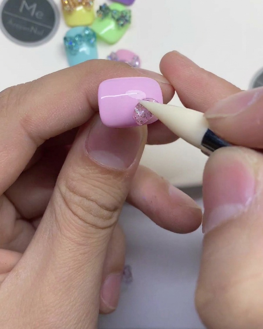Primo piano di una mano che usa uno strumento in silicone bianco per premere un piccolo pezzo di gel glitterato trasparente su ununghia rosa chiaro.