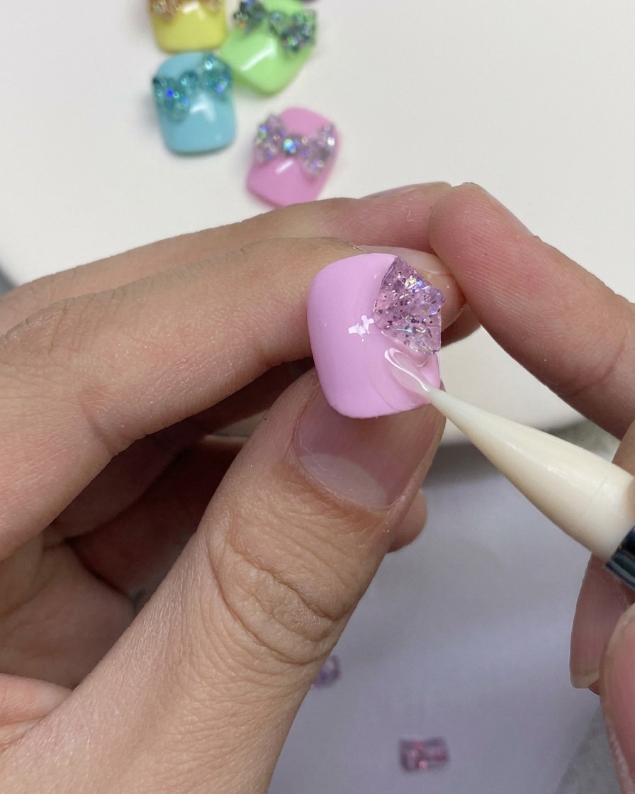 Primo piano di uno strumento in silicone che modella pezzi di gel glitterato nei passanti esterni di un fiocco a farfalla su ununghia rosa.