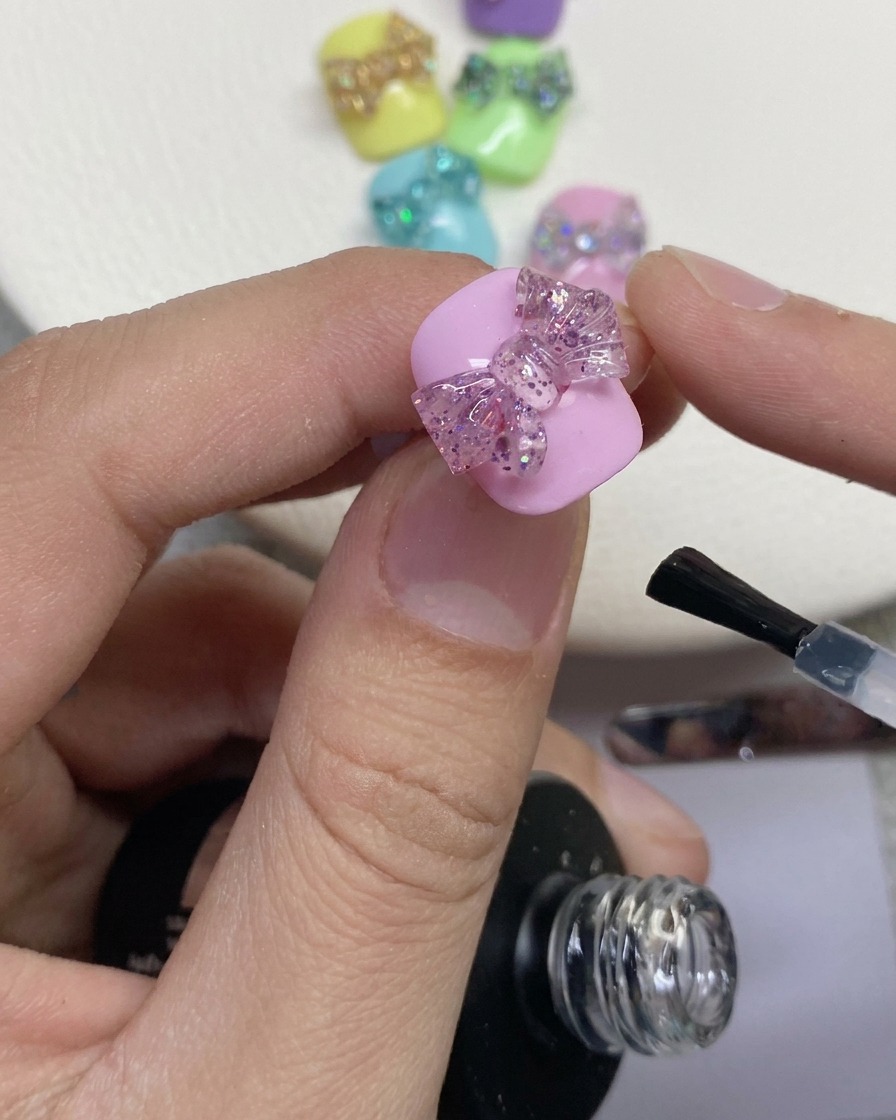 Primo piano di un pennello per unghie che applica il top coat trasparente attorno a un fiocco glitter 3D su ununghia laccata di rosa.