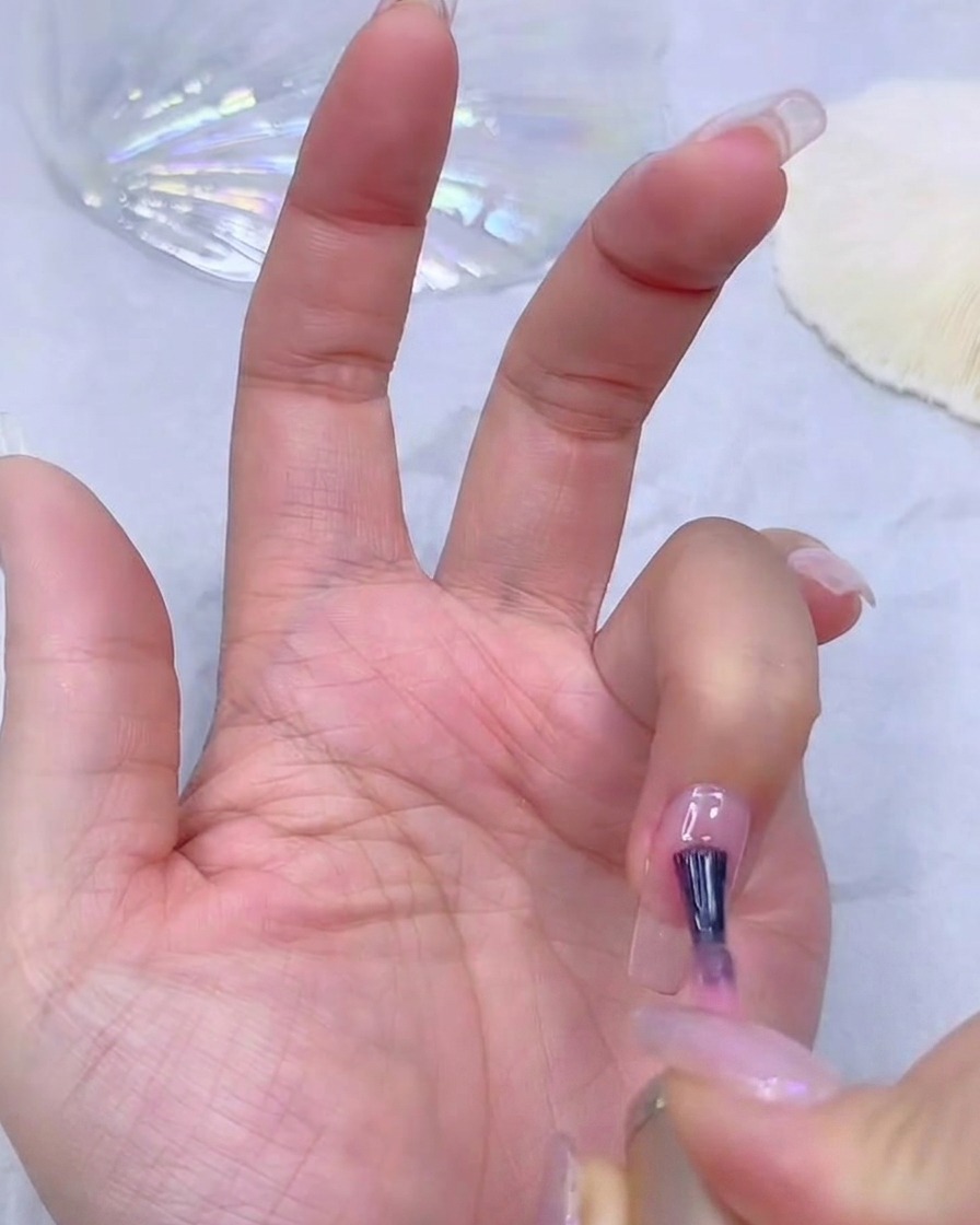 La mano di unonicotecnica che applica una base gel rosa traslucido su una ricostruzione per unghie trasparente.