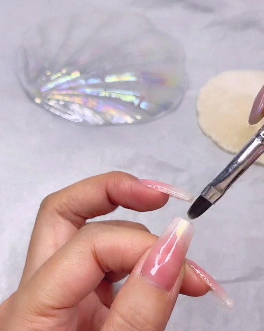 Un pennello fine per unghie che applica un gel trasparente scintillante effetto conchiglia sulla punta di ununghia lunga con sfumatura rosa e bianca.