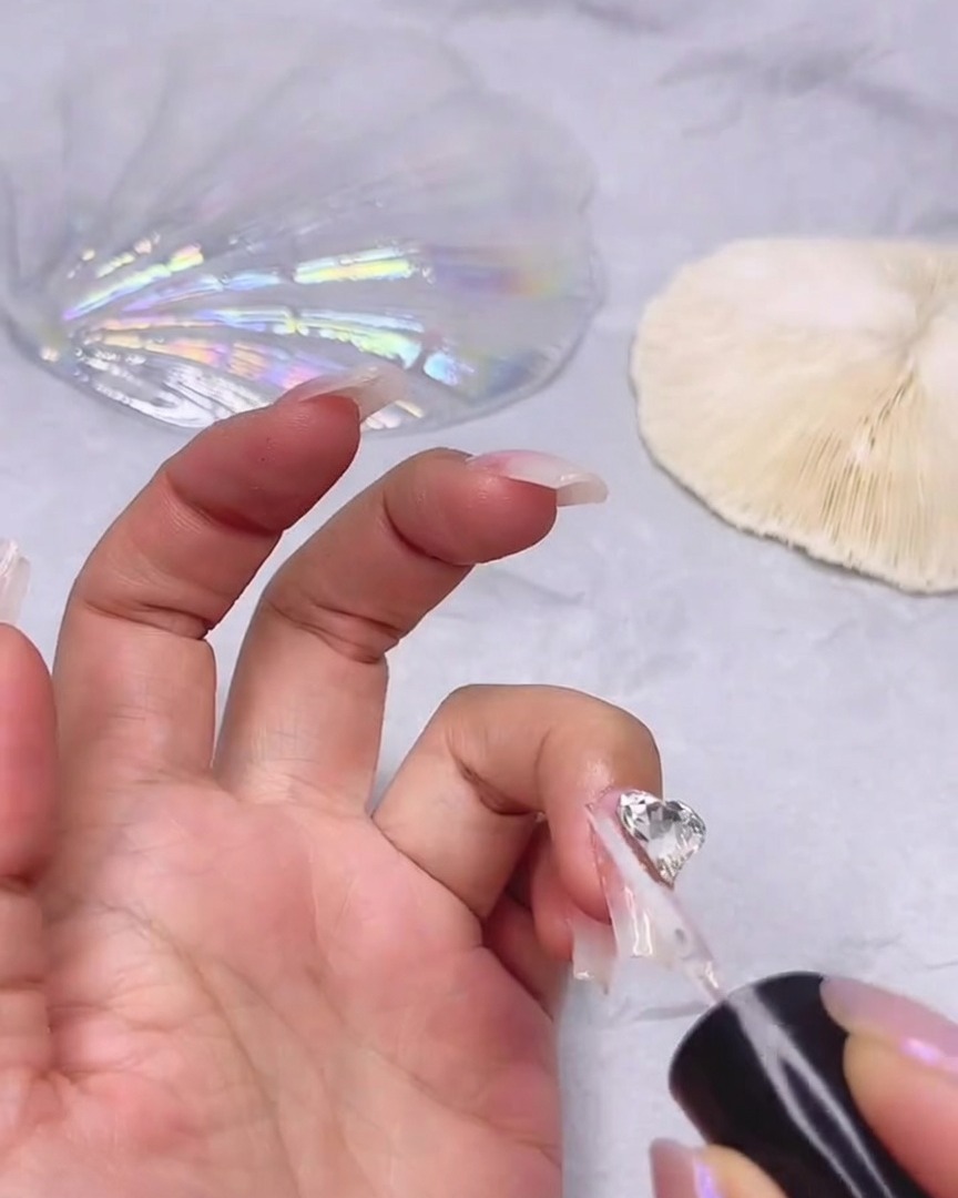 Un pennello fine che applica gel sigillante trasparente attorno ai bordi di un grande strass trasparente a forma di cuore attaccato a ununghia.