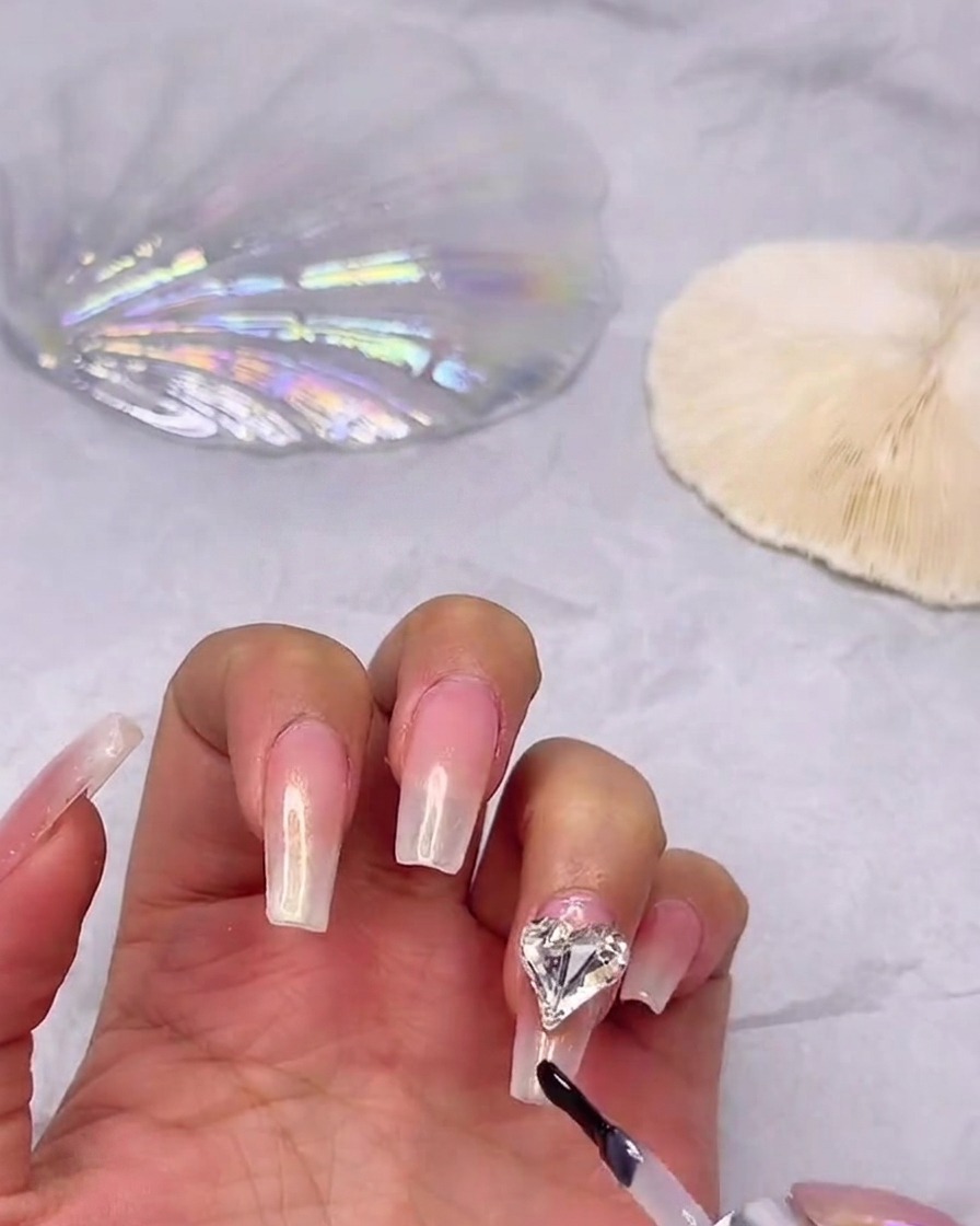 Un pennello per smalto gel che applica un top coat lucido su ununghia sfumata, lavorando con attenzione attorno a uno strass a cuore già fissato.