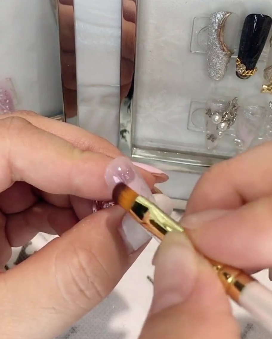 Una mano che tiene un pennello per unghie applicando il gel rosa sulla metà superiore di ununghia a mandorla.
