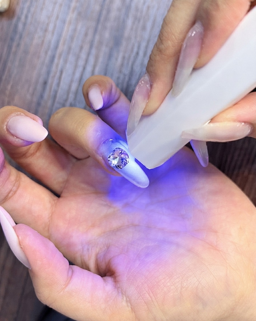 Un primo piano di una mano che raccoglie piccoli strass di cristallo per disporli attorno a una pietra centrale più grande su ununghia curata.
