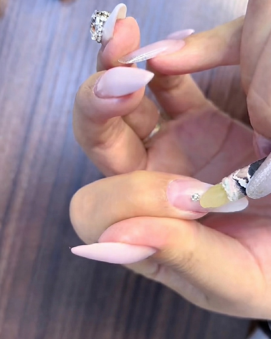 Un pennello sottile che applica gel riempitivo trasparente attorno alla base di un piccolo grappolo di strass su ununghia sfumata.