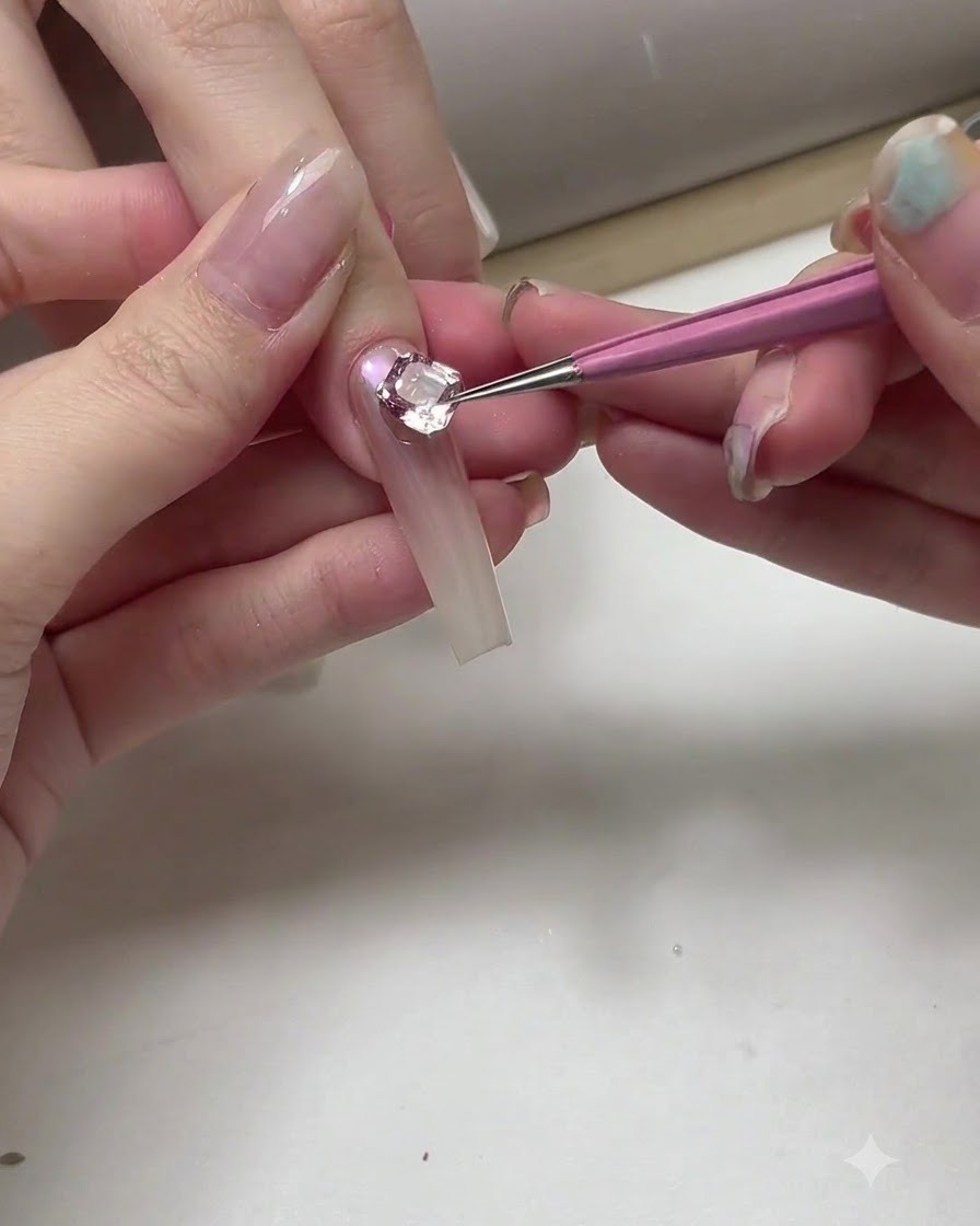 Primo piano di una onicotecnica che usa un pennello sottile e gel argentato per aggiungere delicate linee curve vicino a un grande cristallo su ununghia.