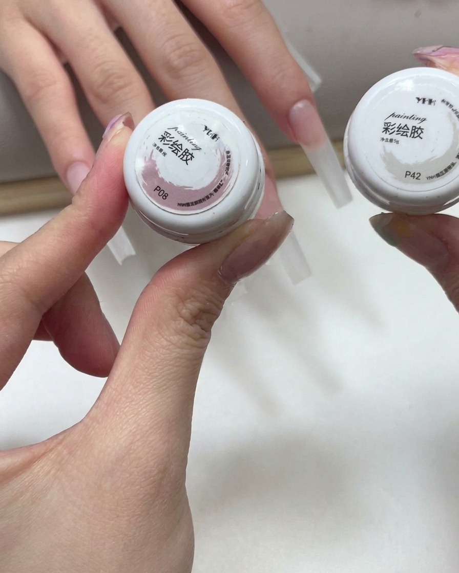 Mani che tengono due piccoli vasetti di gel colorato rosa e bianco con unghie allungate visibili sullo sfondo.