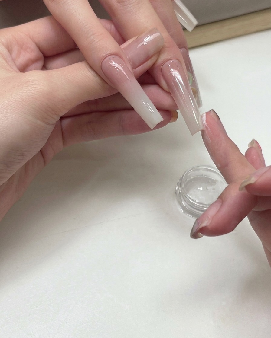 Una onicotecnica che usa la punta delle dita per tamponare polvere di pigmento brillante pesca bianca sulla superficie di ununghia sfumata allungata.
