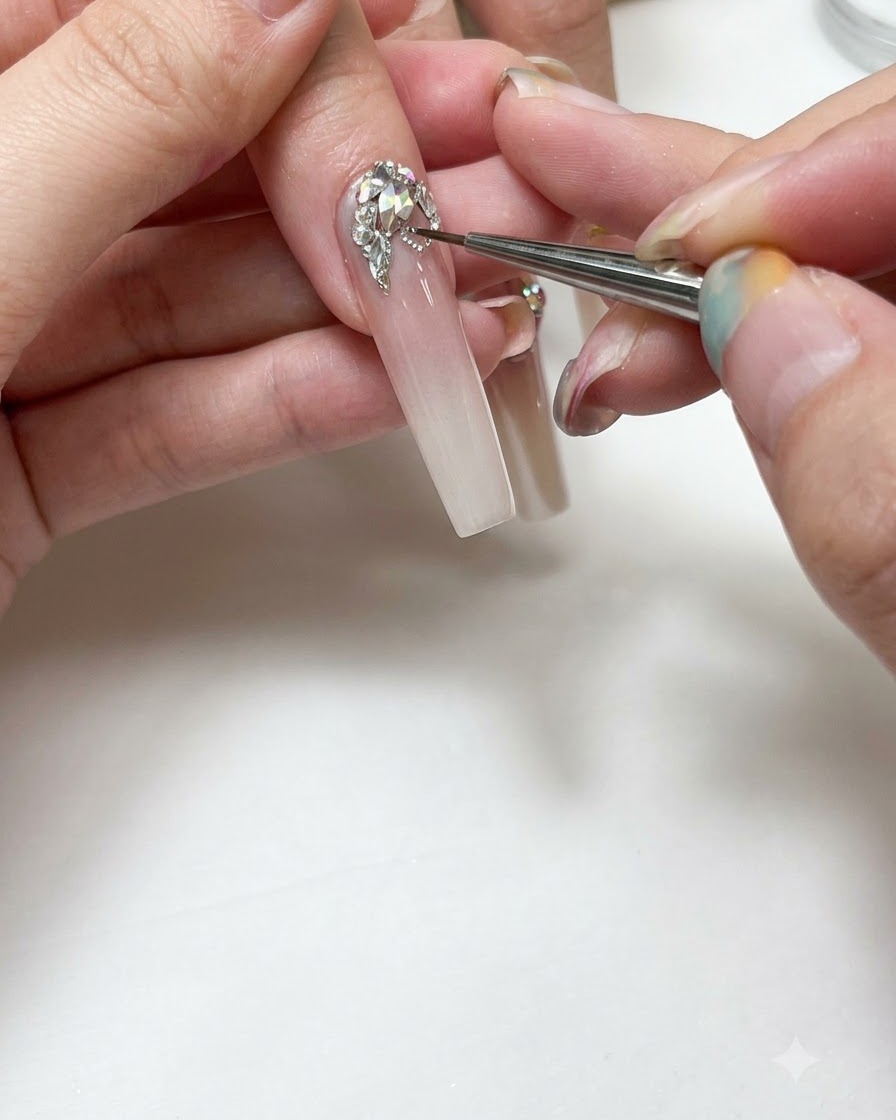 Primo piano di una onicotecnica che usa un pennello di dettaglio fine per dipingere linee in gel argentato attorno a strass trasparenti su ununghia lunga sfumata e allungata.