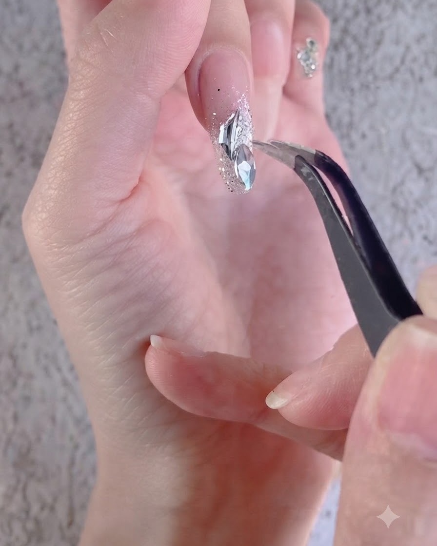 Una mano che utilizza una pinzetta per disporre strass bianchi irregolari su ununghia decorata.