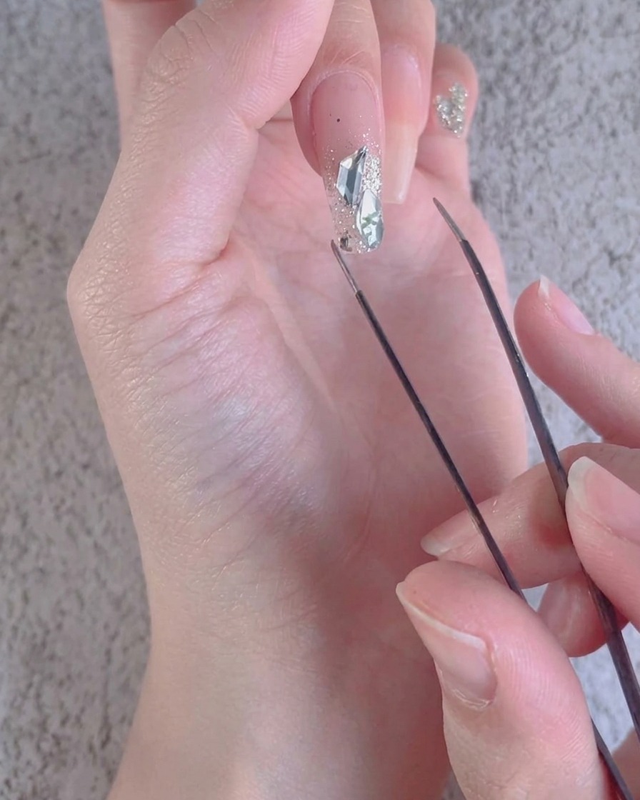 Un primo piano di una pinzetta che regola piccoli accenti di diamante frantumato attorno a pietre più grandi su ununghia.