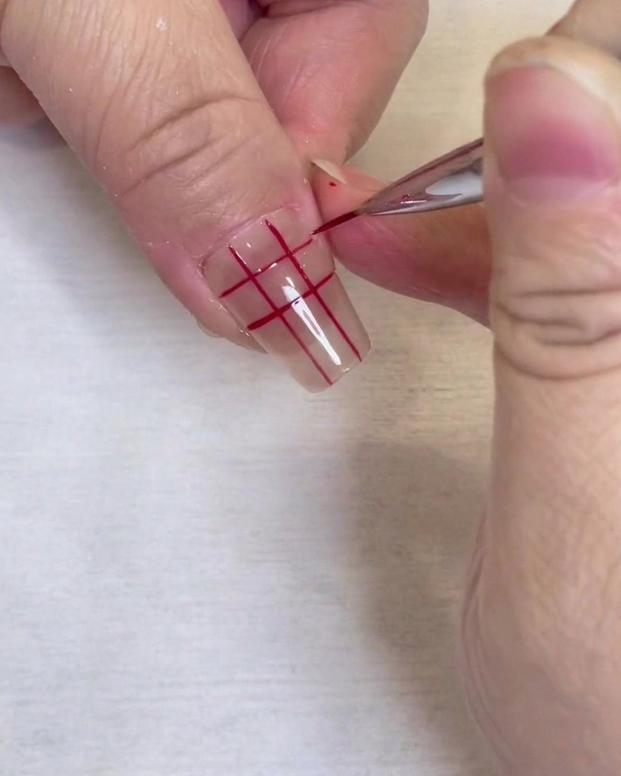 Primo piano di un motivo a griglia rossa completato su ununghia trasparente, tracciato con un pennello liner sottile.