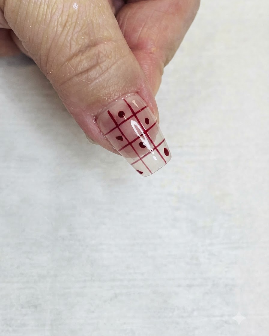 Un pennello liner sottile che riempie i quadratini alternati di un motivo a griglia rossa su ununghia trasparente con smalto rosso.