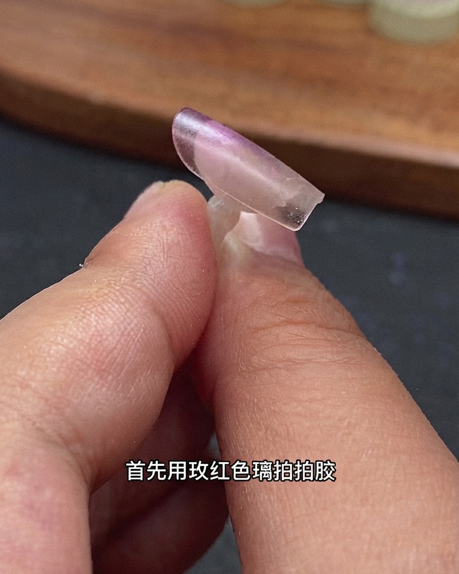 Un primo piano di una tip per ricostrazione unghie trasparente tenuta dalle dita con gel rosa rosso applicato sul bordo posteriore.