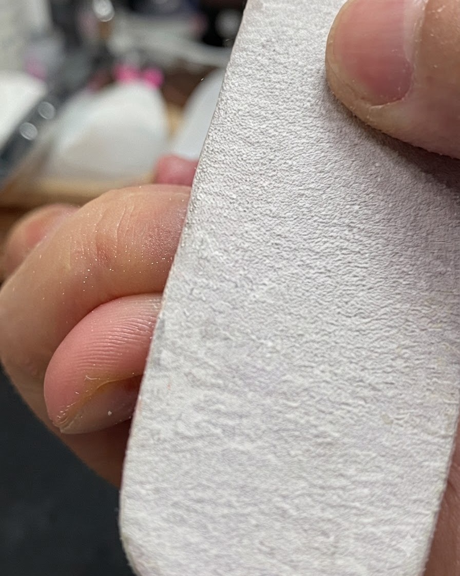 Una mano che tiene un blocco buffer bianco mentre lima leggermente la superficie di ununghia naturale.