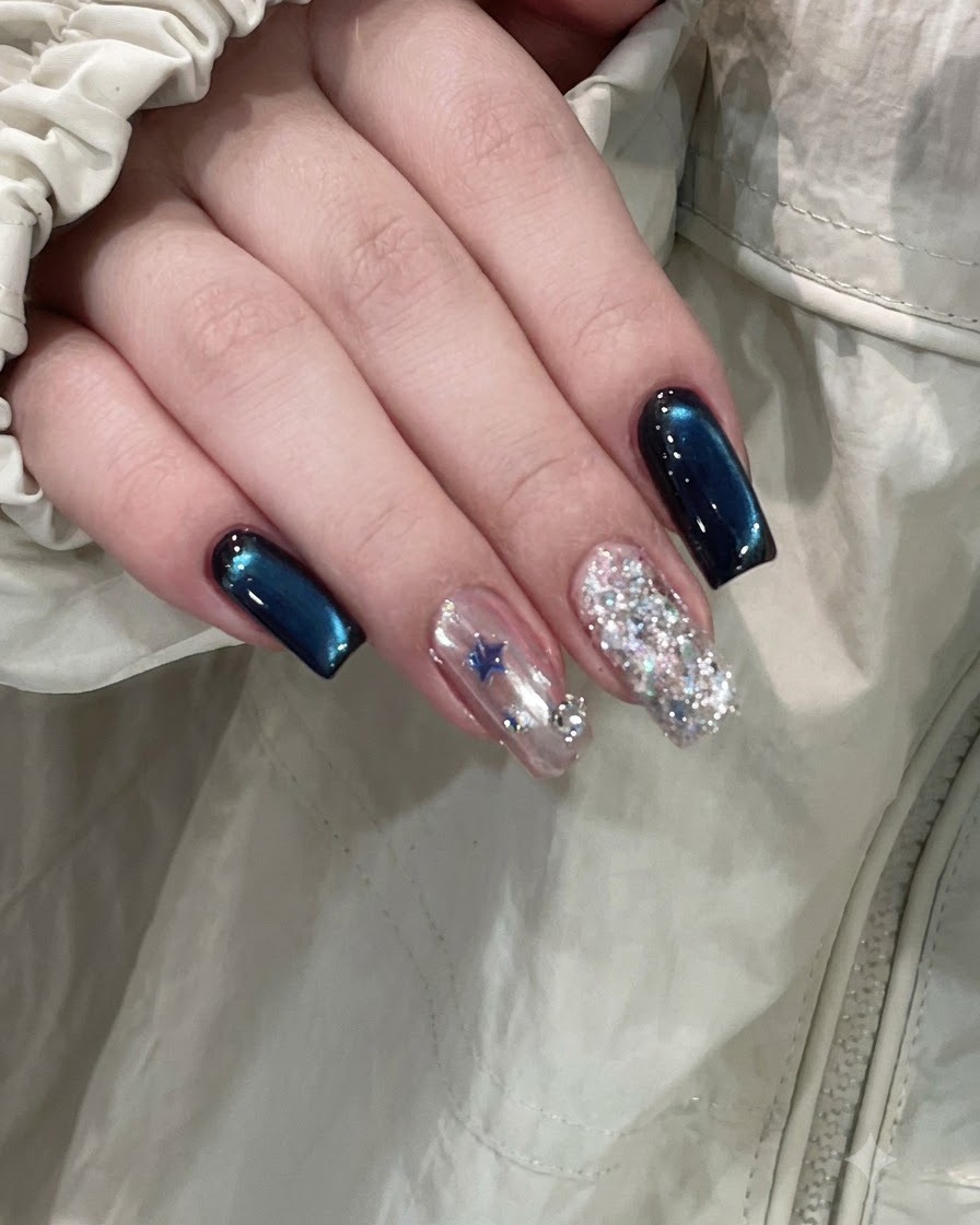 Una mano finita che mostra unghie cat-eye blu profondo accanto a ununghia daccento decorata con stelle e ununghia interamente glitterata.