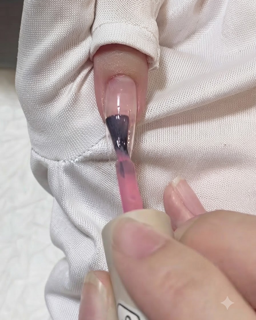 Un pennello che applica una pennellata di gel rosa traslucido lungo il centro di una tip per unghie trasparente.