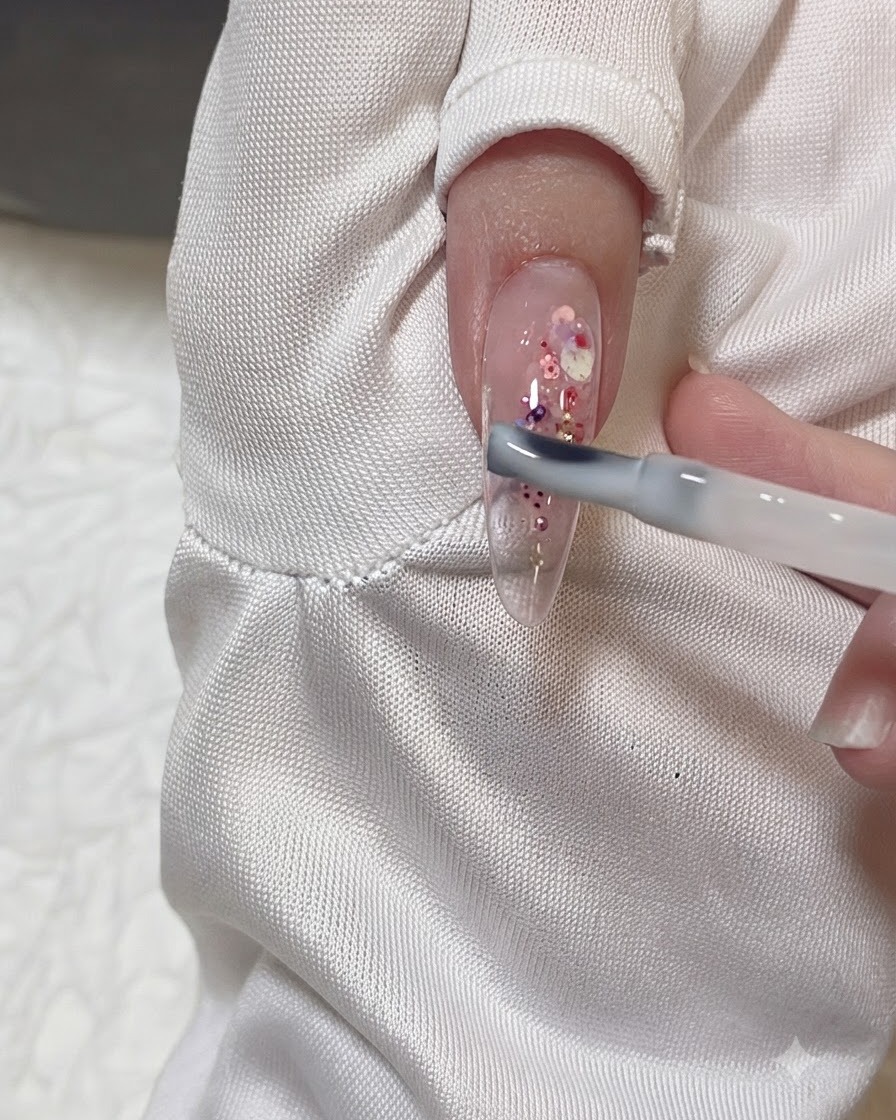Un pennello da gel che applica uno strato di gel lattiginoso trasparente su ununghia a mandorla trasparente decorata con scaglie di glitter rosa e viola.