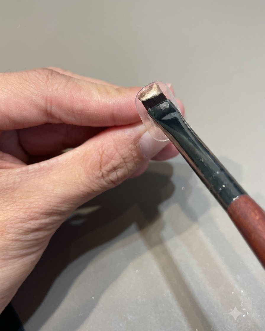 Un pennello carico di smalto gel cat eye rosa oro tenuto sopra un supporto trasparente per esposizione unghie.