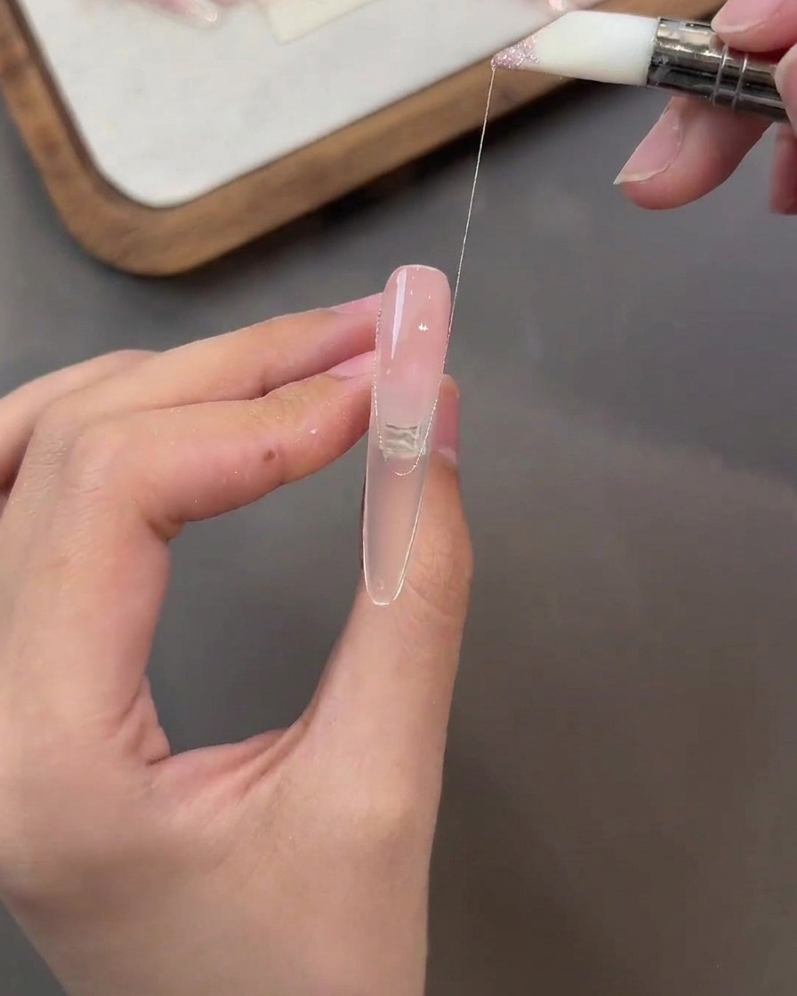 Un pennello che stende un sottile filo di gel rosa glitterato sulla superficie di ununghia color nude.
