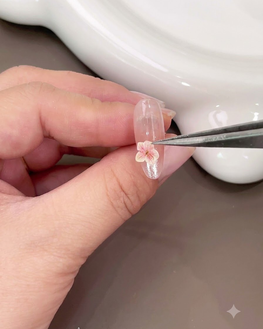Un paio di pinzette che posizionano un piccolo fiore secco rosa sulla parte inferiore di ununghia lucida.