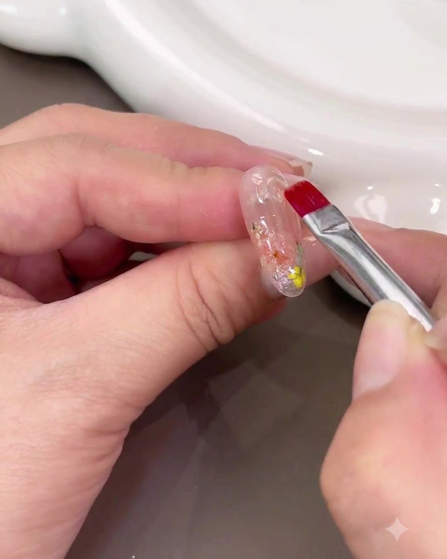Un pennello per unghie che applica uno strato spesso di gel costruttore trasparente sulla nail art con fiori secchi.