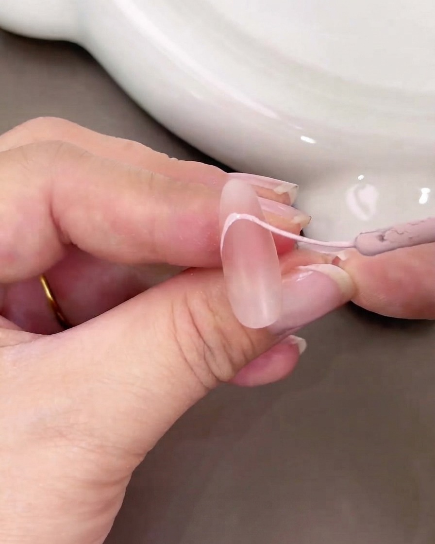 Primo piano di una mano che usa un pennello sottile per dipingere una curva French tip rosa chiaro su ununghia nude.